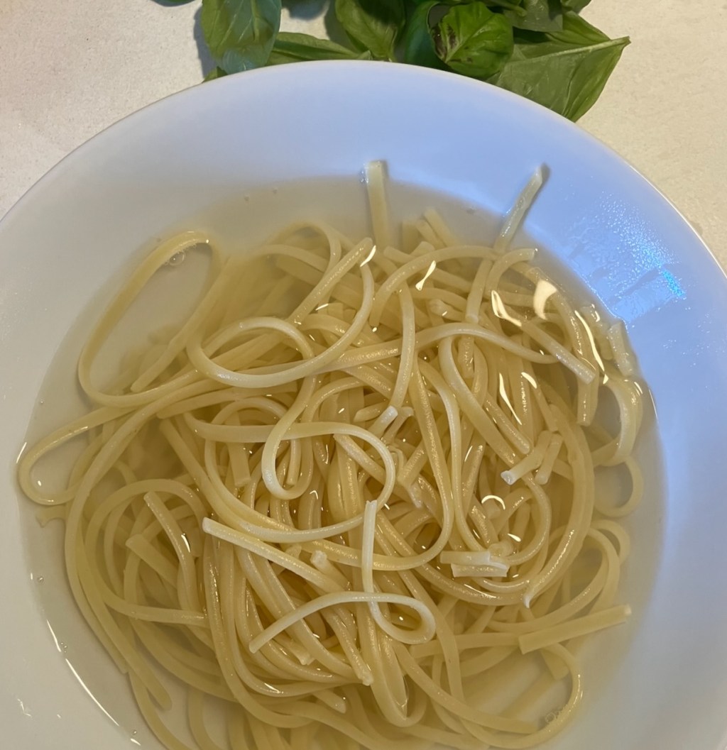 Linguine in Kochwasser in einer weissen Schale, dahinter Basilikum